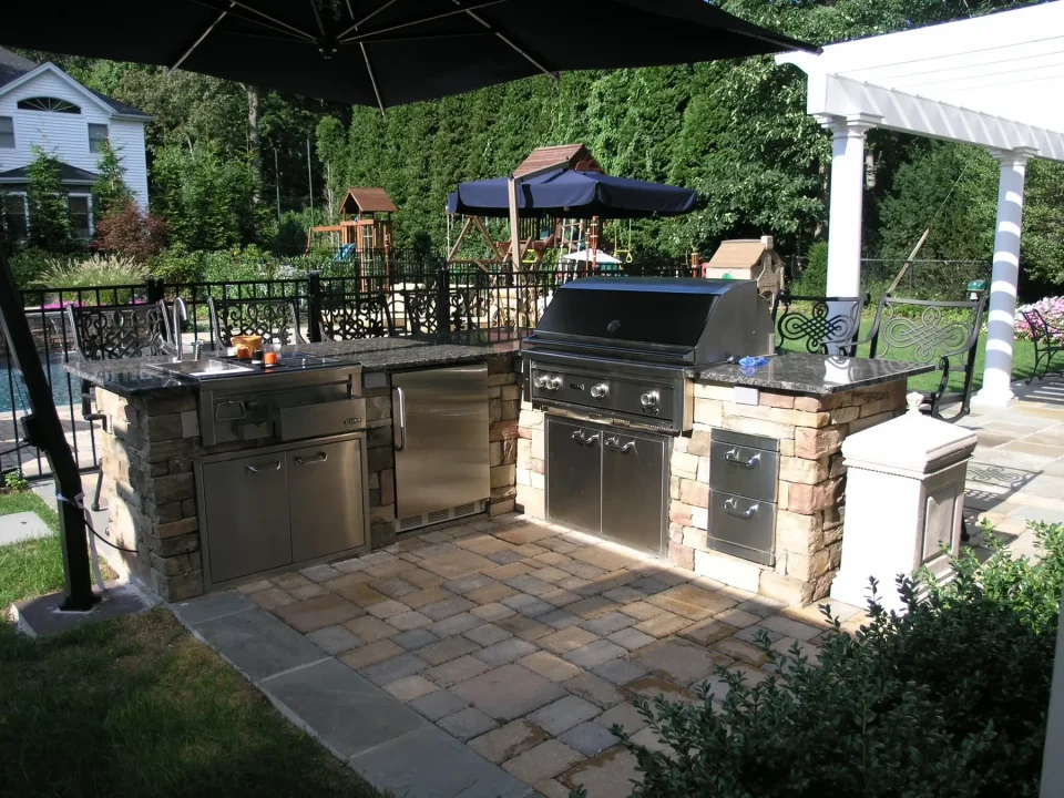Outdoor kitchen with grill, fridge and sink on stone patio, fenced garden backdrop.