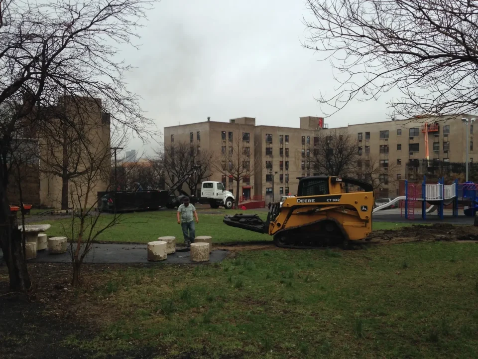 A muddy park with a person, skid steer loader, trucks, and apartment buildings in the background.