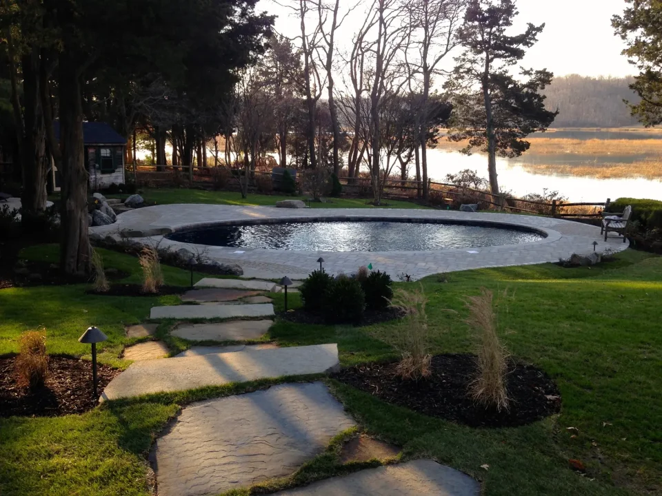 Oval pool with stone path and bench by a lake at sunset.