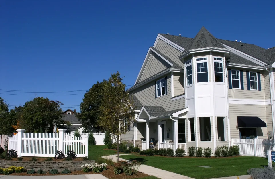 Two-story beige house with white trim, a white picket fence, and a clear blue sky.