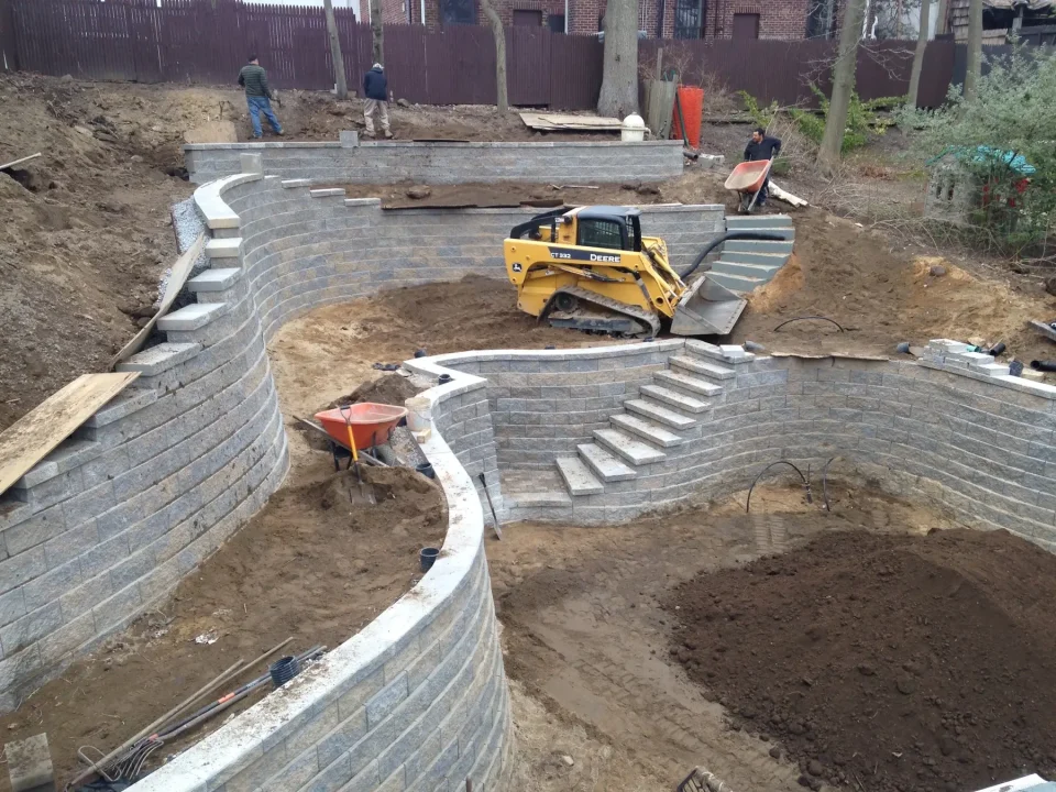 Construction site with terraced retaining walls, stairs in progress, and workers operating machinery.