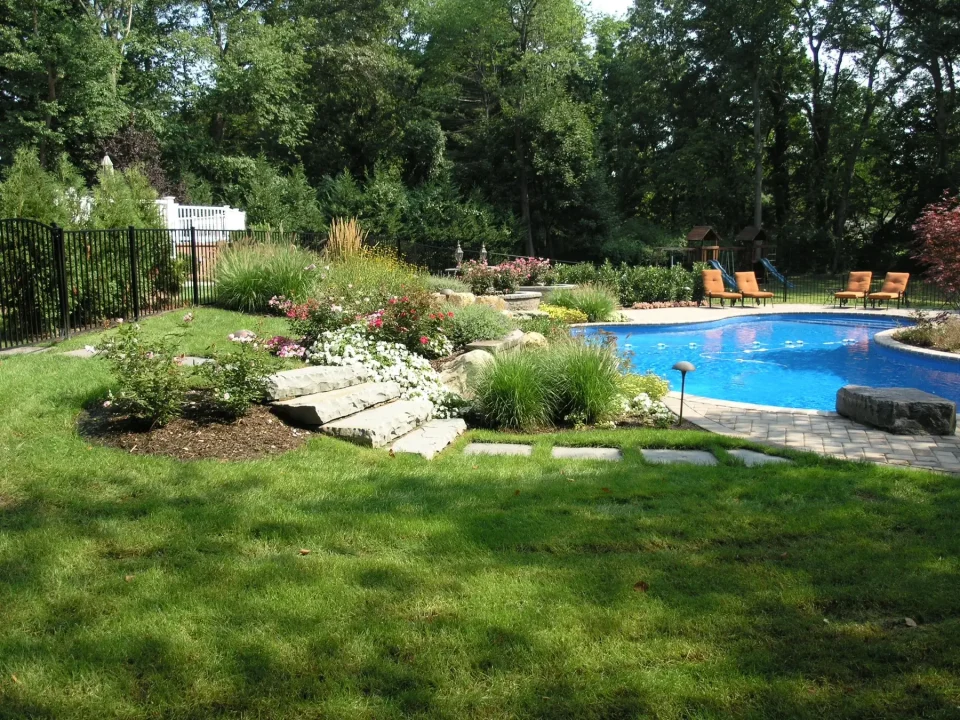 Backyard with a swimming pool, landscaped garden, and patio chairs on a sunny day.