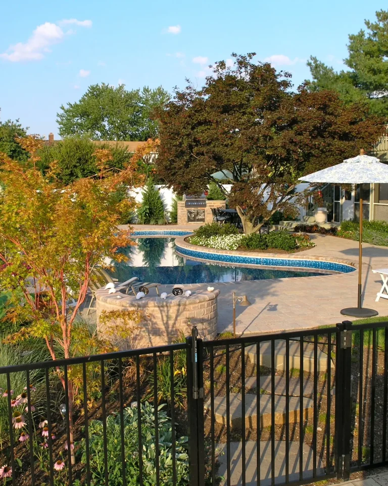 Lush garden with a curvy pool, umbrella-shaded table, and a stone fire pit.