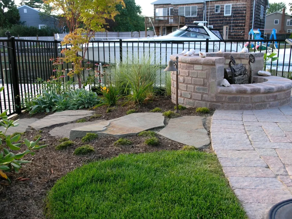 A backyard garden with stepping stones leading to a brick fire pit, a pool behind a metal fence, and a boat.