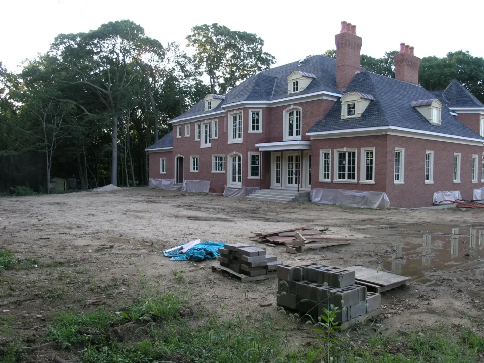 A large, unfinished red brick house with multiple chimneys and construction materials in the front yard.