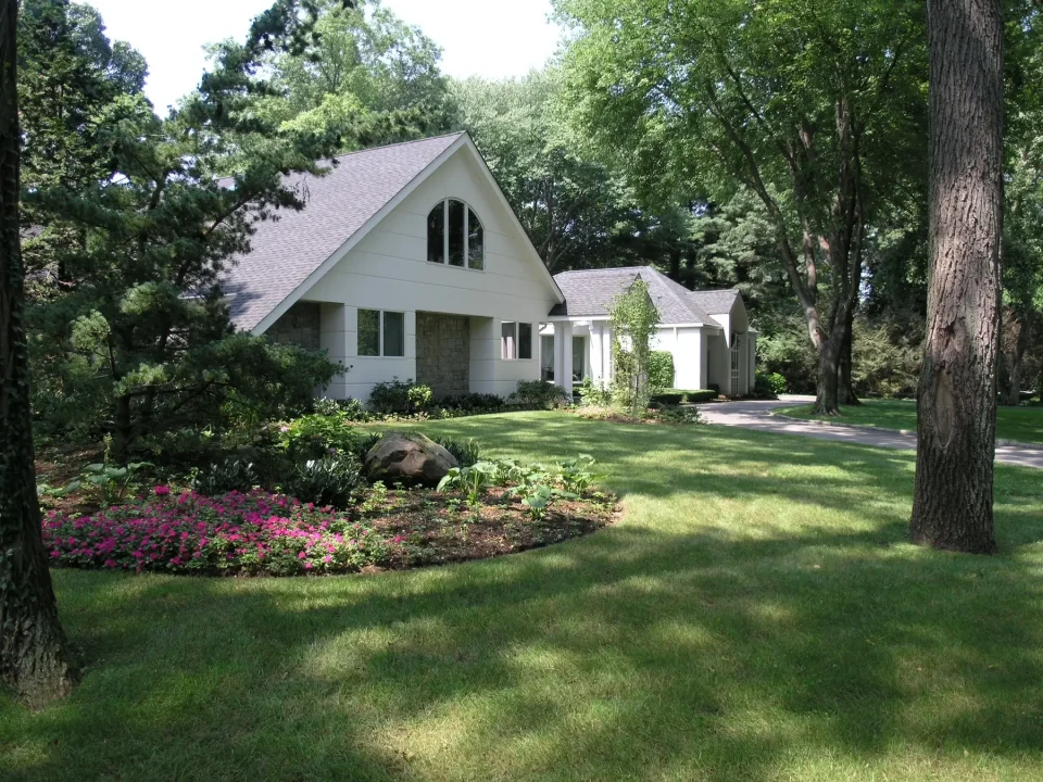 White house with large windows nestled among green trees and landscaped garden.
