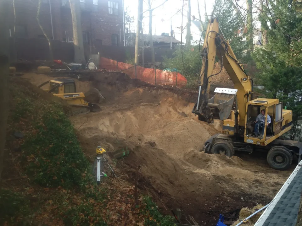 Two excavators operating on a construction site with a dirt pile and trees in the background.