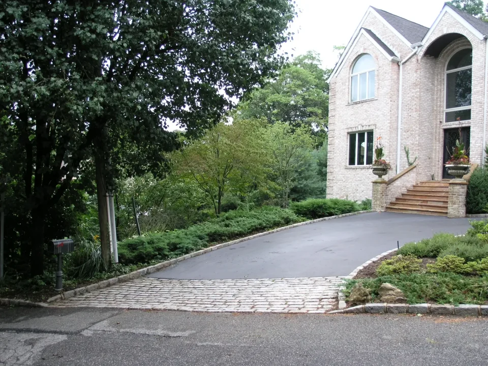Driveway leading to a two-story brick house with tall windows and front steps, surrounded by trees and shrubs.