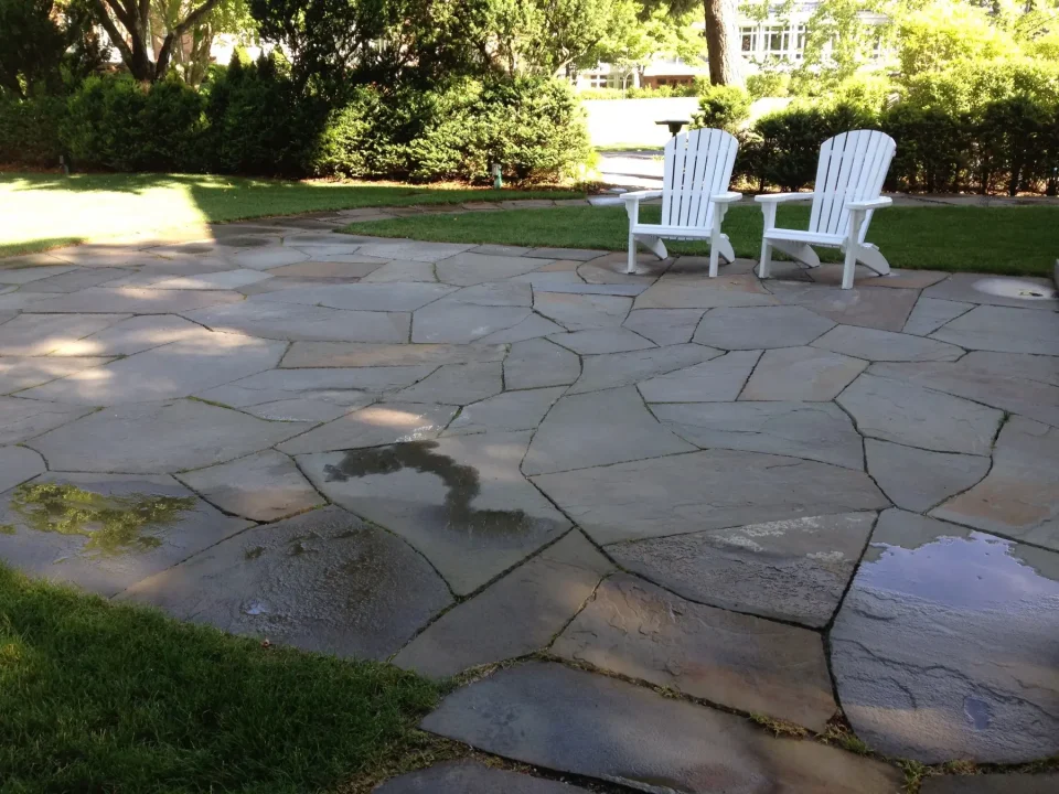 Two white Adirondack chairs on a wet stone patio with surrounding greenery.