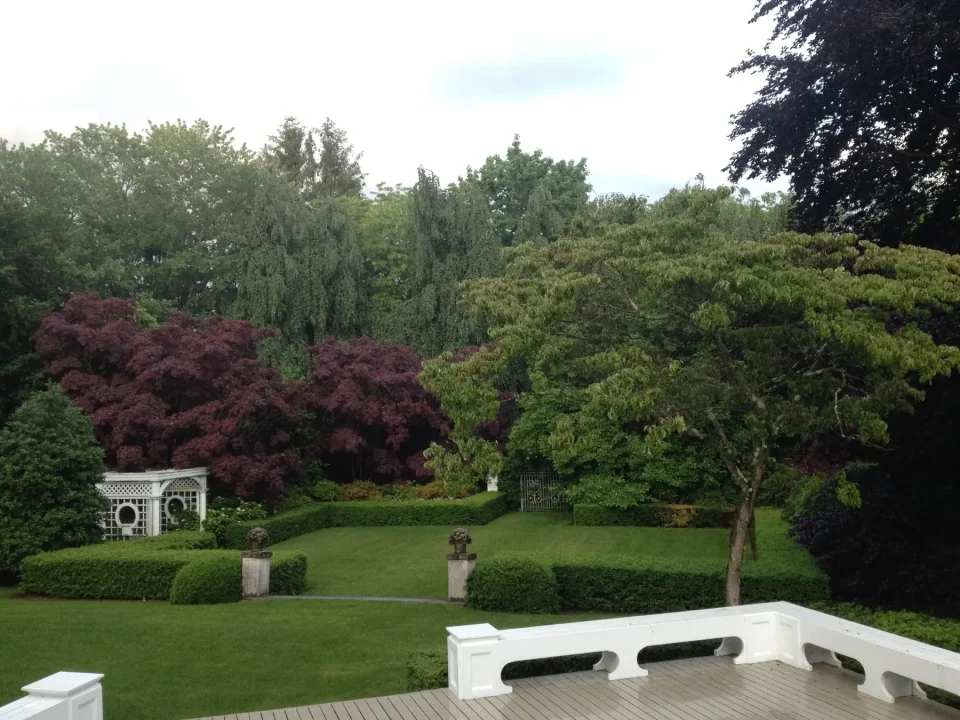Lush garden with purple trees, a gazebo, trimmed hedges, and a white railing in the foreground.