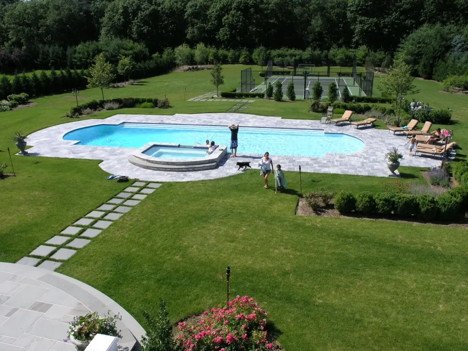 Aerial view of a backyard with a blue pool, hot tub, sun loungers, and a tennis court. People and a dog are present.