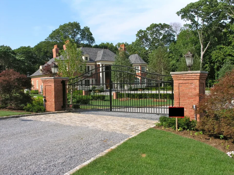 Elegant brick entrance with wrought iron gates leading to a grand estate, amidst lush greenery.