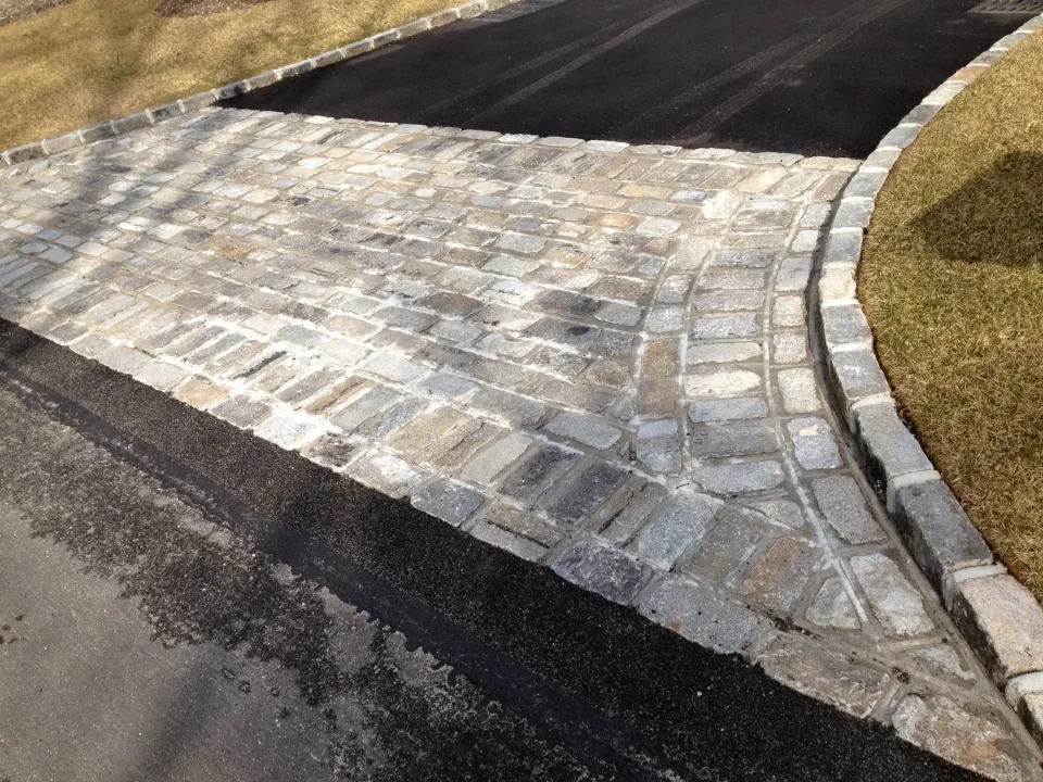 A cobblestone driveway intersection with asphalt road and curbs.