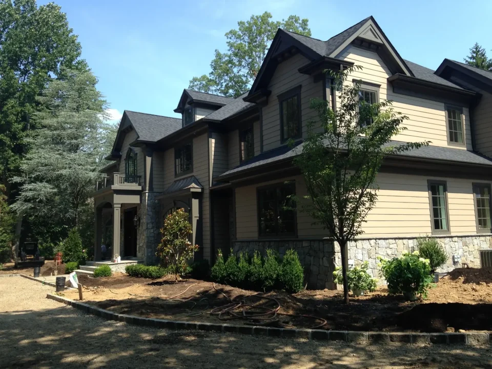 Two-story house with a gray roof and beige siding, surrounded by trees and under construction landscaping.