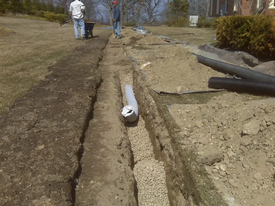 Two people working on a trench with drain pipes, in a residential area.