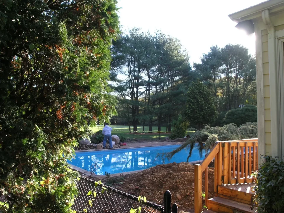 A backyard with a pool and person working, surrounded by trees, with a wooden deck in the foreground.
