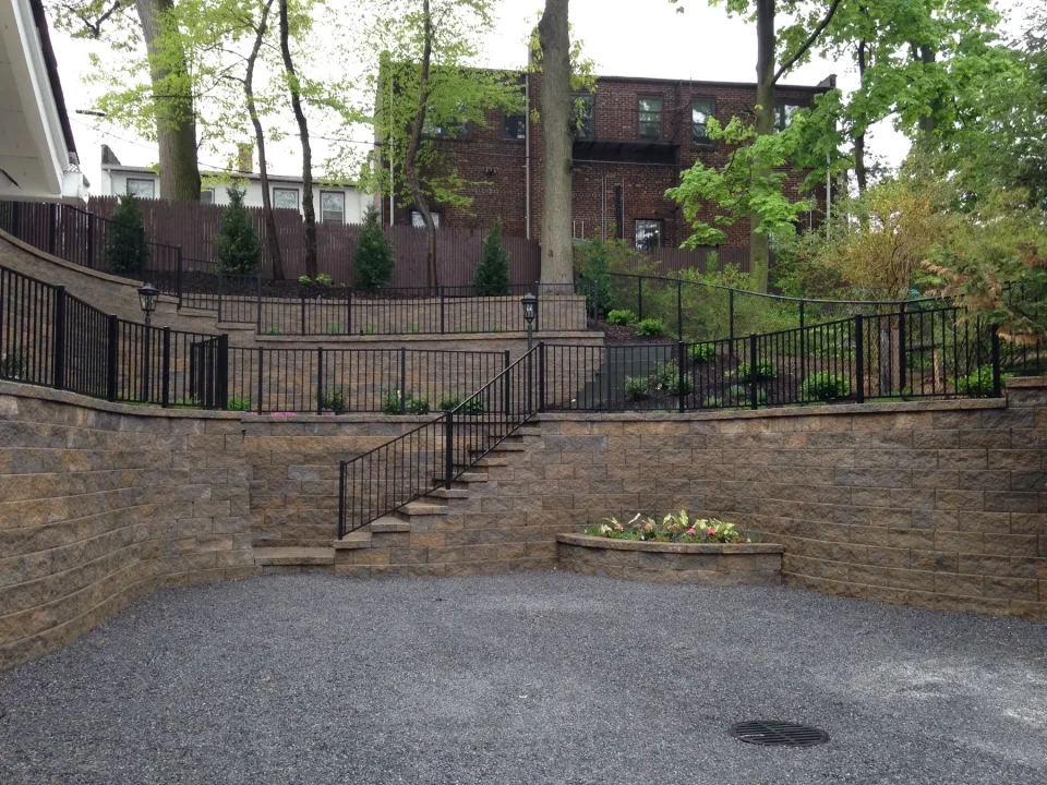 Stone-walled courtyard with steps and iron railing, surrounded by greenery and a brick building.