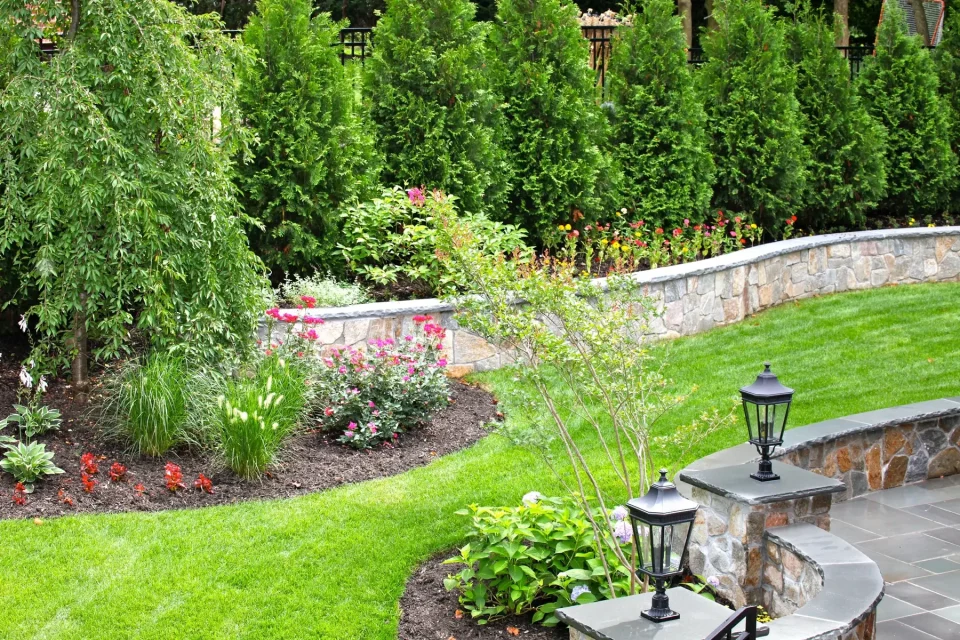 Well-manicured garden with stone wall, flowering plants, and classic lanterns on a patio.