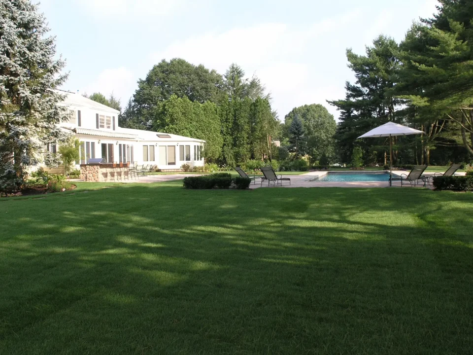 Lush backyard with a white house, swimming pool, sun chairs, and an umbrella.