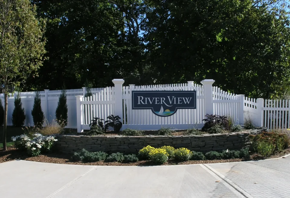 White picket fence with "River View" sign, landscaped garden, and trees in the background.