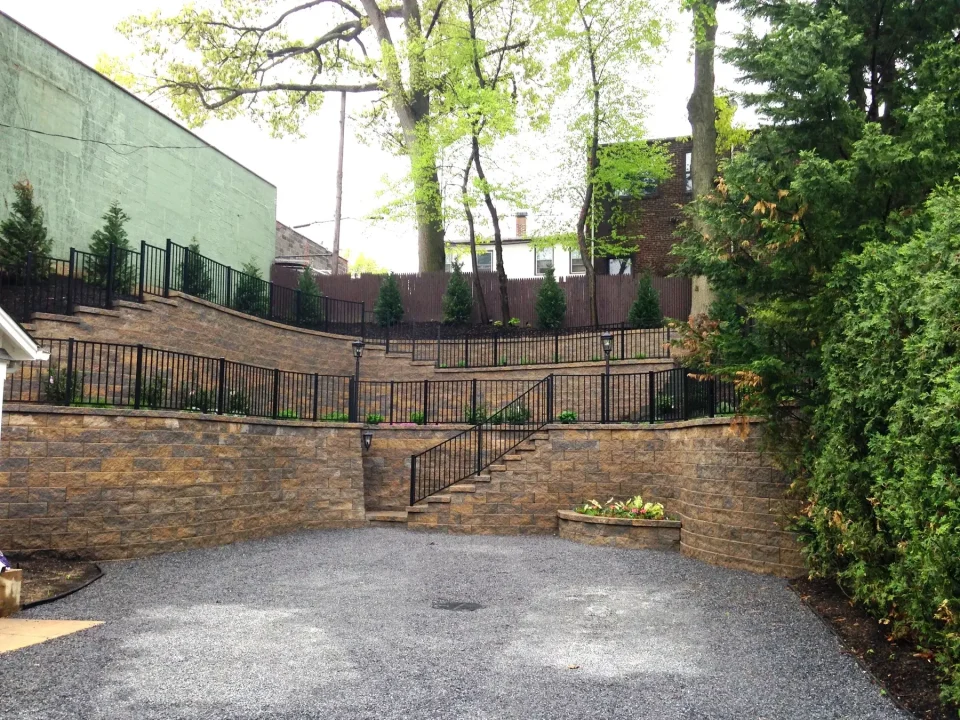 Driveway leading to tiered retaining walls with stairs, railings, and greenery on a cloudy day.