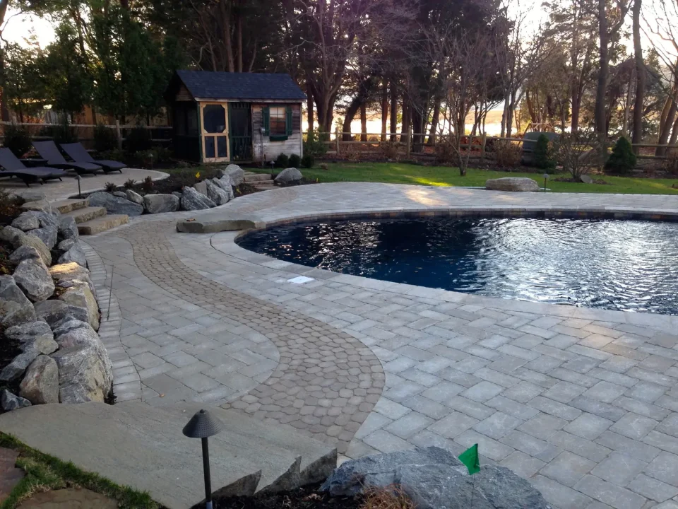 Backyard with a curved pool, stone pathways, and a small house, with trees in the background.