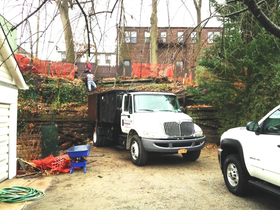 A work truck parked beside a pile of debris with a person standing on top, near residential buildings.
