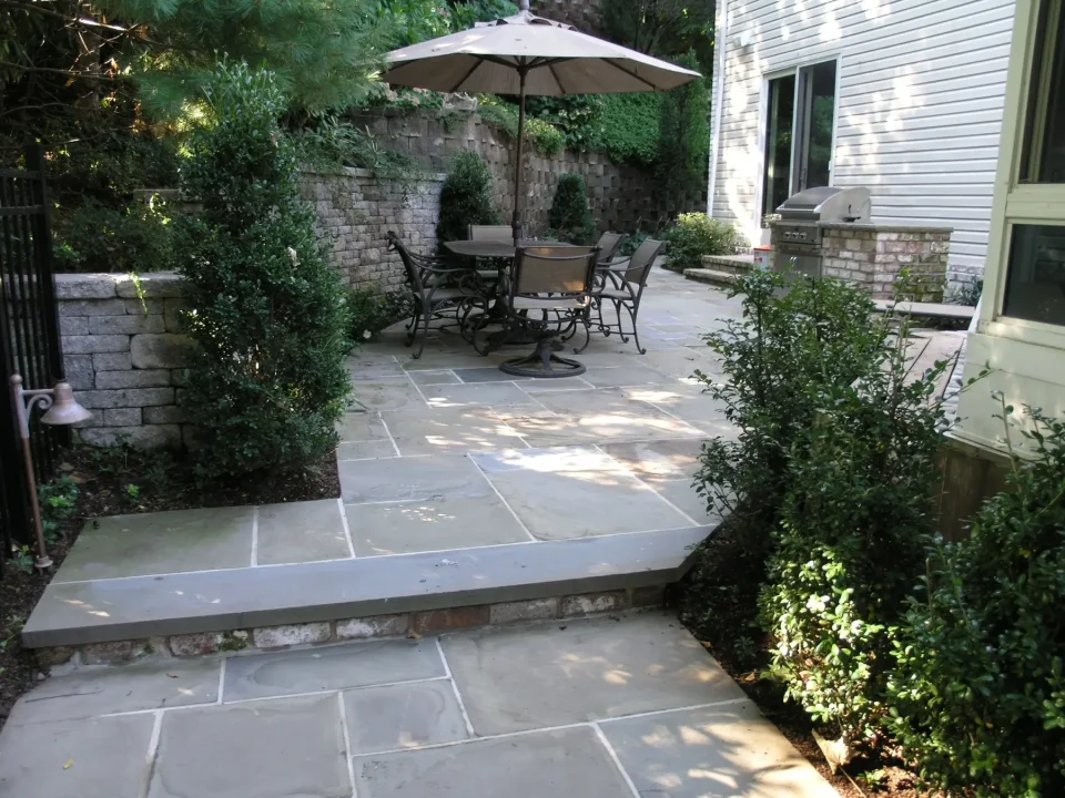 A stone patio with outdoor furniture under an umbrella, adjacent to a house and garden.