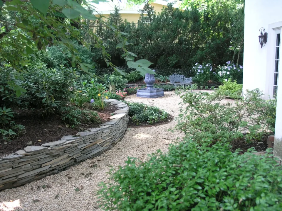 A tranquil garden with a pebble path, lush greenery, and a stone wall.