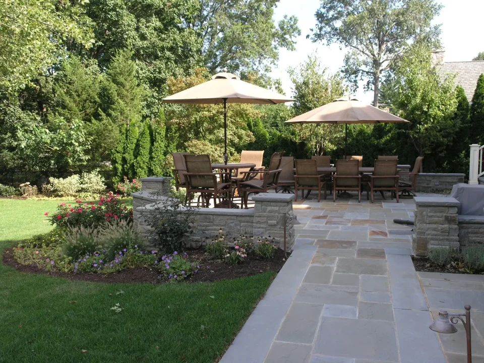 Elegant outdoor patio area with dining furniture under umbrellas, surrounded by lush greenery and flowers.