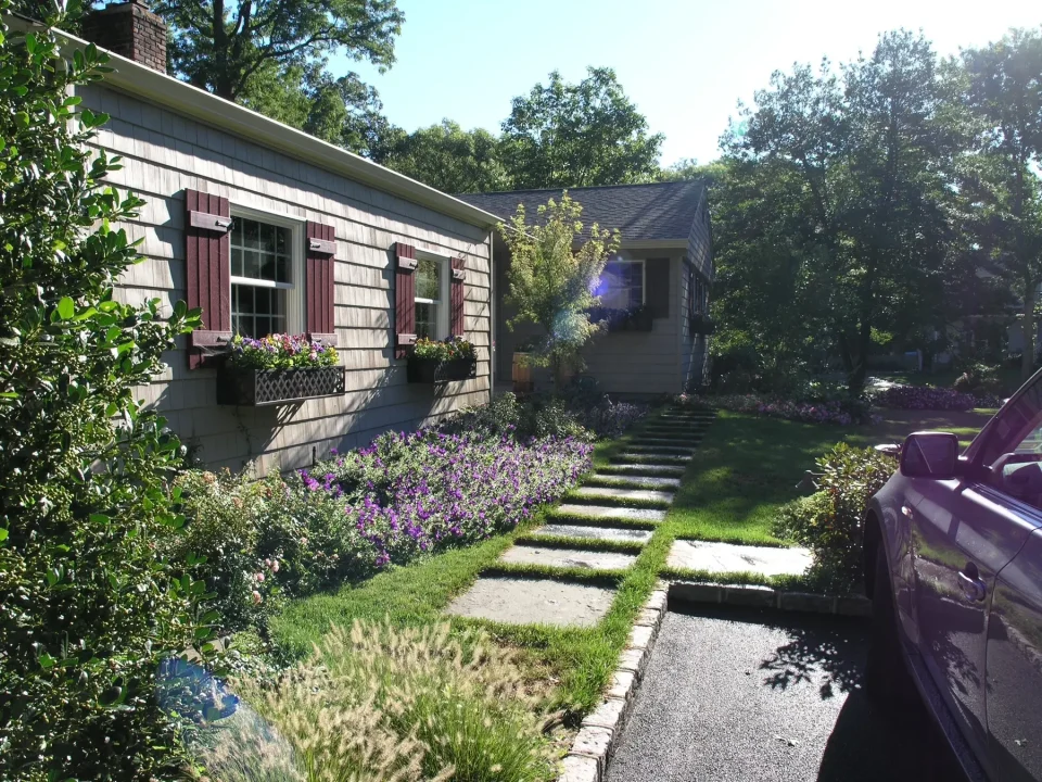 Suburban house with flower boxes, a stone path, lush landscaping, and partial car view.