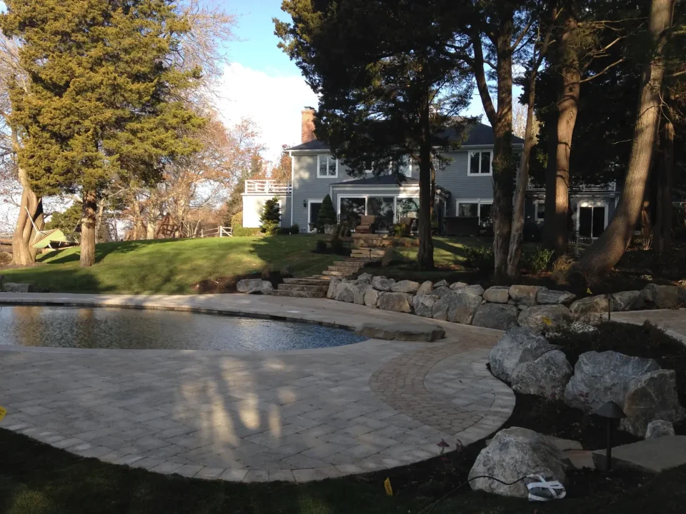 Backyard with a pool, patio, and a two-story house surrounded by trees and rocks.
