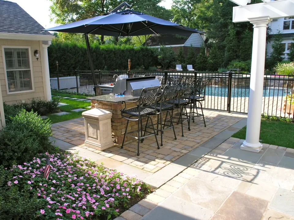 Outdoor patio with grill, dining set, umbrella, and a view of a pool and garden.