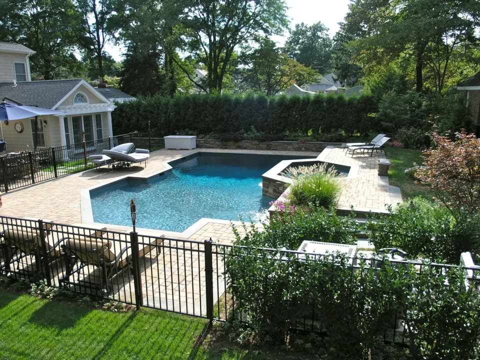 Backyard with a blue swimming pool, sun loungers, surrounded by greenery and a house.