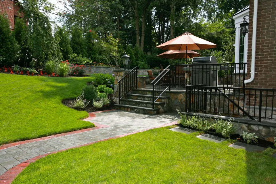 Lush garden with stone steps, patio, umbrella, and iron railing beside a brick house.