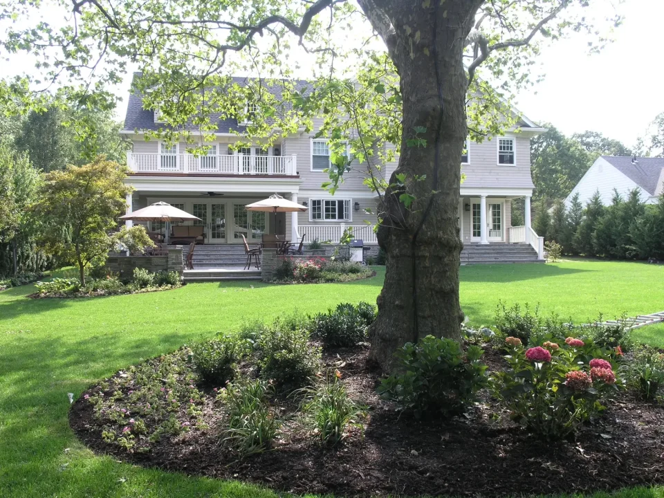 White two-story house with a porch, umbrellas, and landscaped garden with a large tree.