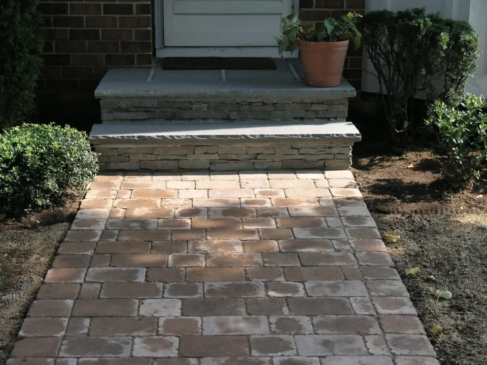 Brick walkway leading to a stone doorstep with a white door, flanked by green shrubs.