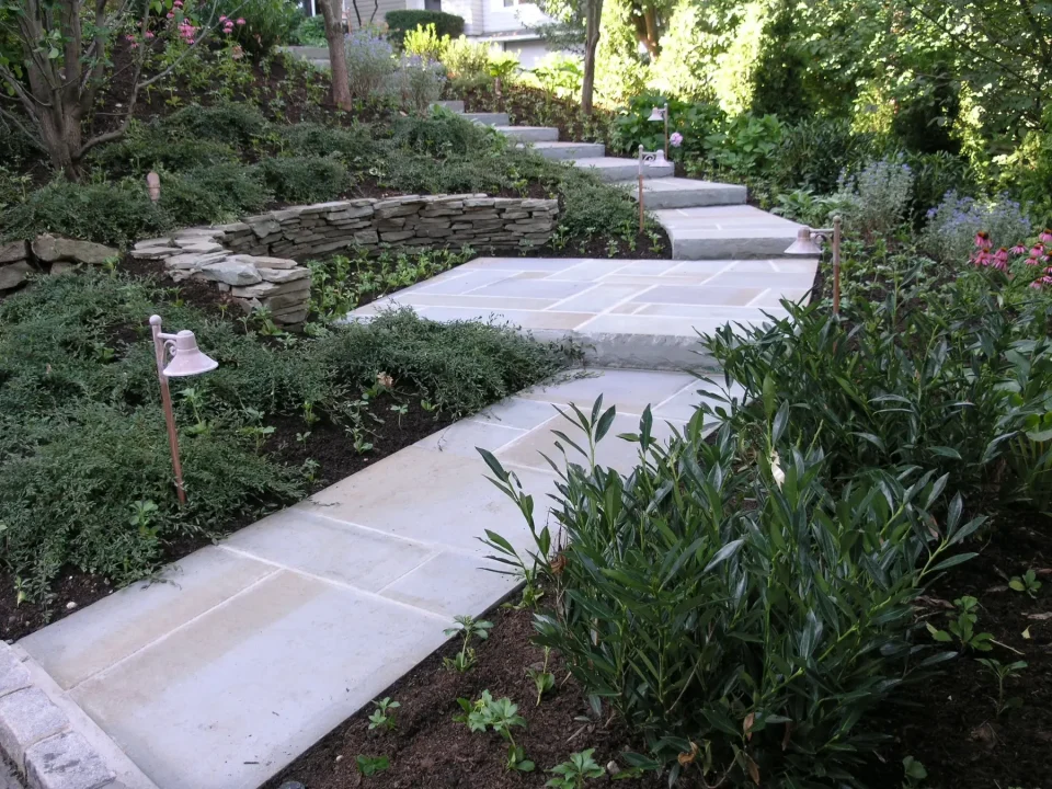 Stone pathway with green plants and a small bell on the side in a garden setting.