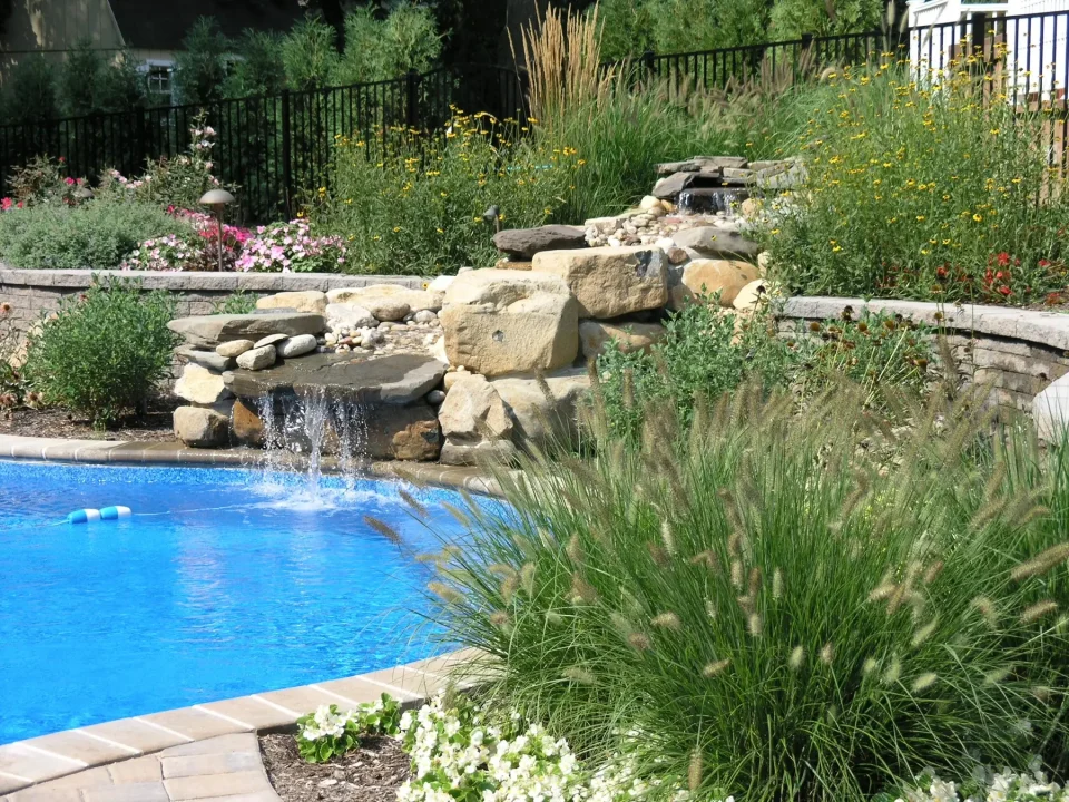 A landscaped backyard with a pool and cascading waterfall feature, surrounded by flowering plants and ornamental grasses.