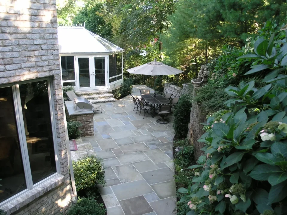 Stone patio with furniture next to a house and sunroom, surrounded by greenery.
