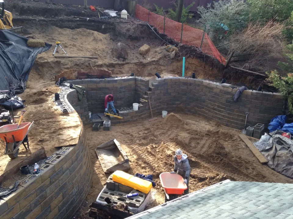 Construction of a curved brick wall in an excavated site with building materials and workers.