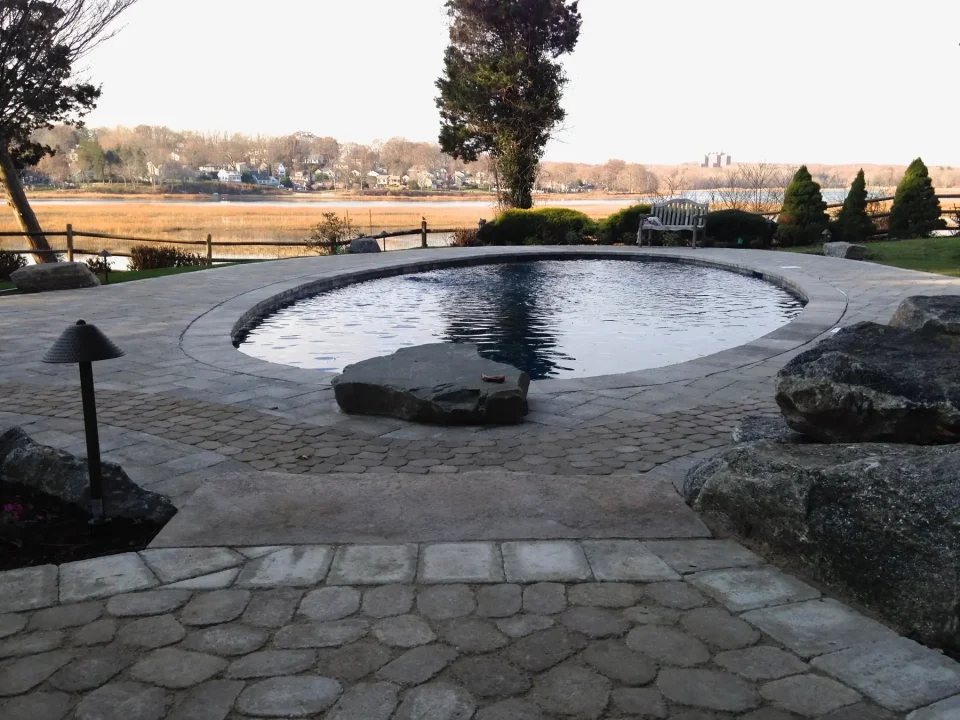 A circular outdoor pool surrounded by paving and rocks, with a river and trees in the background.