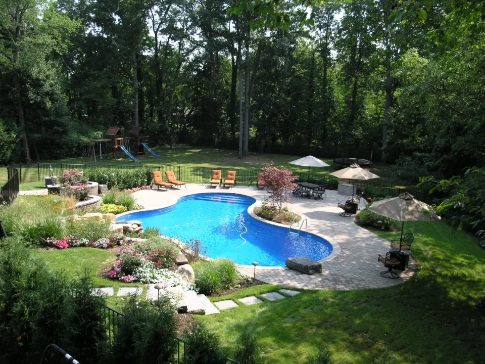 A beautifully landscaped backyard with a curvy pool, patio area, sun umbrellas, and a playground surrounded by trees.