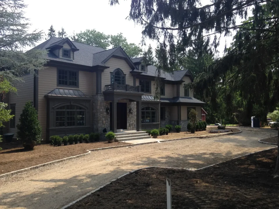 Two-story house with stone foundation and gray siding, surrounded by trees and a dirt driveway.