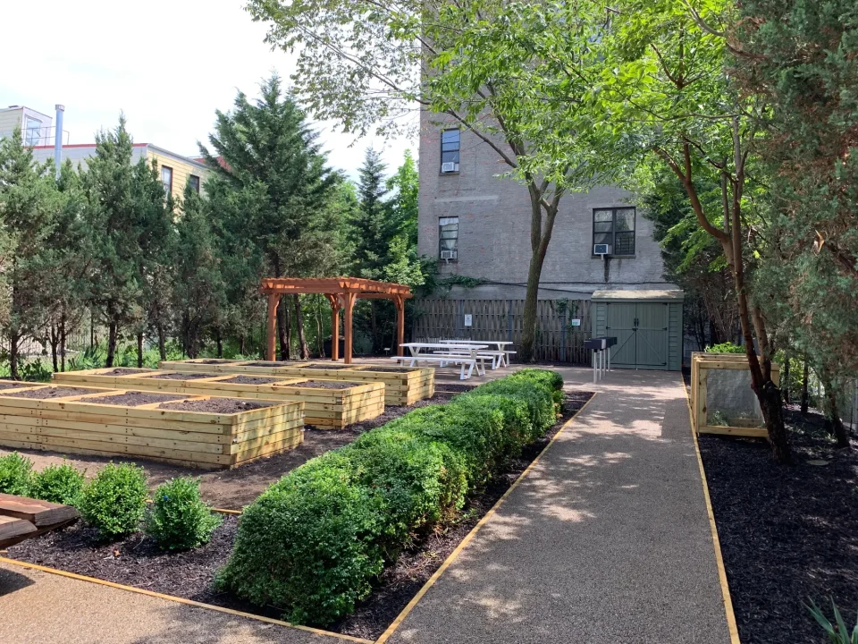 Urban garden with raised beds, a pergola, and a seating area, surrounded by trees and buildings.