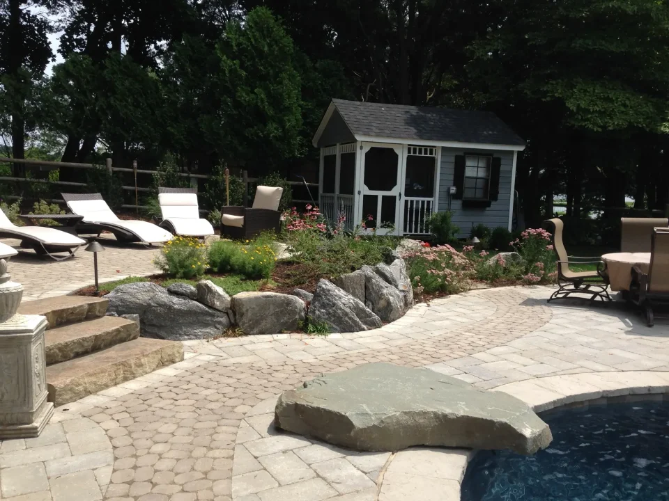 Backyard with pool, stone paving, lounge chairs, and a small gray garden shed surrounded by greenery and flowers.